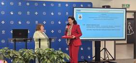 Pokaż zdjęcie: Sala konferencyjne. Przed dużym ekranem z nazwą konferencji stoją dwie kobiety, przemawiają do mikrofonów.