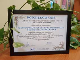 Pokaż zdjęcie: W czarnej ramce stoją podziękowania od dyrektora Łódzkiego Oddziału Wojewódzkiego NFZ dla Łódzkiego Oddziału PFRON. W tle widać zielone liście. 