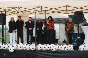 Pokaż zdjęcie: Scena plenerowa. Na scenie stoją kobiet i mężczyźni elegancko ubrani, przemawiają do mikrofonu.