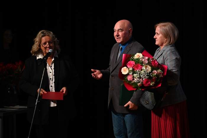 Pokaż zdjęcie: Na scenie stoją trzy osoby, dwie kobiety blondynki oraz mężczyzna w garniturze. Jedna z kobiet trzyma bukiet kwiatów. 