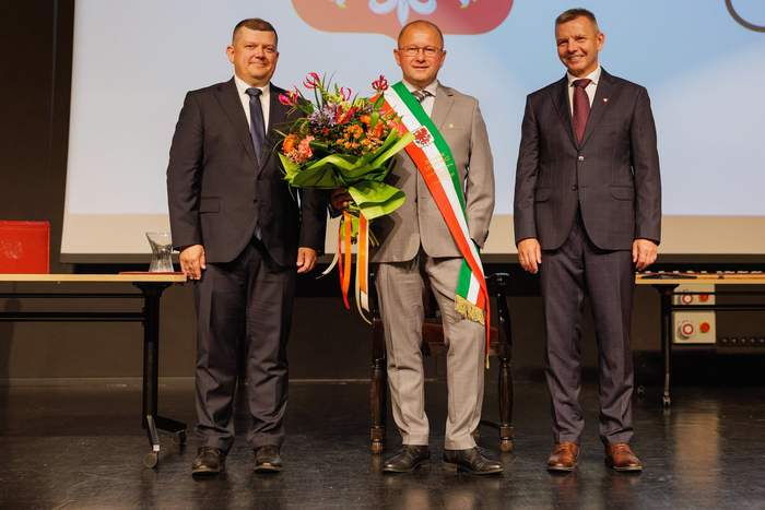 Pokaż zdjęcie: Trzech mężczyzn ubranych w garnitury stoi obok siebie, a w środku jeden z nich trzyma kwiaty i ma na sobie szarfę w kolorach Miasta Gorzowa Wielkopolskiego