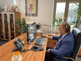 Pokaż zdjęcie: Przy biurku, na fotelu siedzi mężczyzna ubrany w białą koszulę i niebieską marynarkę. Na głowie ma słuchawki, wpatruje w monitor. W tle okno z kwiatami doniczkowymi stojącymi na parapecie.