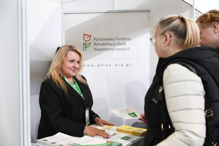 Stoisko Państwowego Funduszu Rehabilitacji Osób Niepełnosprawnych. Kobieta za ladą rozmawia z uczestniczką targów i przekazuje materiały informacyjne.