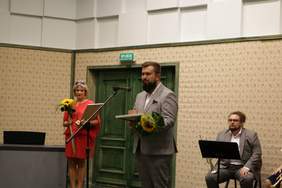 Pokaż zdjęcie: Przed mikrofonem stoi mężczyzną słusznej postury, ubrany elegancko w jasny garnitur w kratę. Trzyma dyplom oraz bukiet kwiatów. Z tyłu stoi kobieta w czerwonej sukience oraz  siedzi mężczyzna w garniturze, na twarzy ma okulary, trzyma trąbkę.