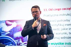 Przedstawiciel PFRON podczas wystąpienia na konferencji „Biznes i Inkluzja”. Prelegent przemawia do publiczności, trzymając mikrofon i pilot do prezentacji. W tle wyświetlane są slajdy dotyczące wsparcia osób z niepełnosprawnościami.