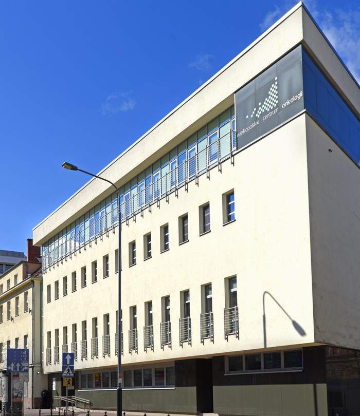 Pokaż zdjęcie: Trzypiętrowy budynek w kolorze kremowym z dużą liczbą okien. Na górze po prawej stronie znajduje się napis: „Wielkopolskie Centrum Onkologii”. W tle niebo, które jest niemal całkowicie bezchmurne.