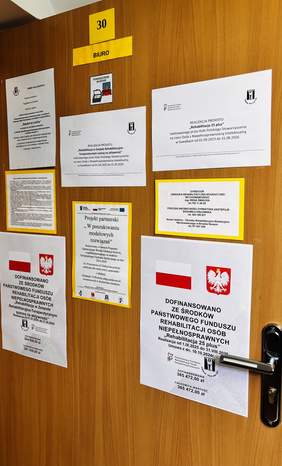 Pokaż zdjęcie: Drzwi oznaczone numerem 30 z napisem „Biuro”; na drzwiach umieszczone są tablice informujące o realizacji projektów dofinansowanych ze środków PFRON, w tym programu „Rehabilitacja 25 plus”.