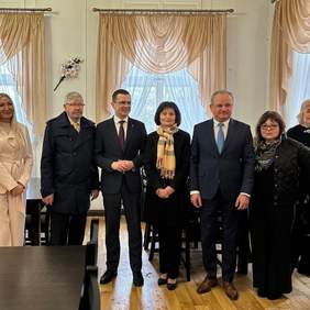 Pokaż zdjęcie: W pokoju stoi grupa kilku osób. Są w niej kobiety i mężczyźni. Wszyscy są elegancko ubrani. W tle widać trzy okna. 