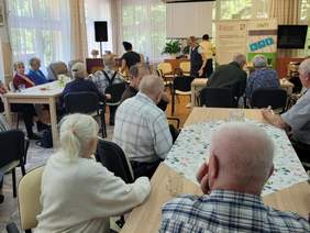 Pokaż zdjęcie: Duża sala w Centrum Seniora. Na krzesełkach siedzi liczna grupa seniorów - kobiety i mężczyźni. W tle osoby które opowiadają o programach PFRON. Widać duże zainteresowanie wśród słuchaczy,