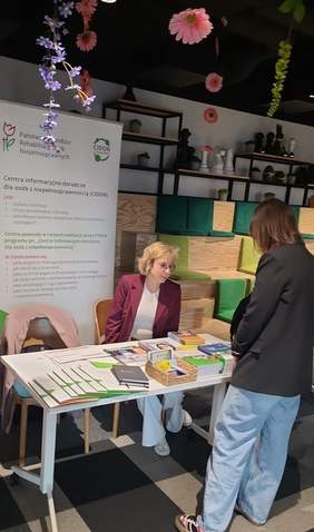 Pokaż zdjęcie: Pracownik PFRON rozmawia z uczestniczką targów przy stoisku informacyjnym z ulotkami. W tle stoi baner z logotypami „Państwowego Funduszu Rehabilitacji Osób Niepełnosprawnych”, a także „Centra informacyjno-doradcze dla osób z niepełnosprawnościami” zwanym CIDON.