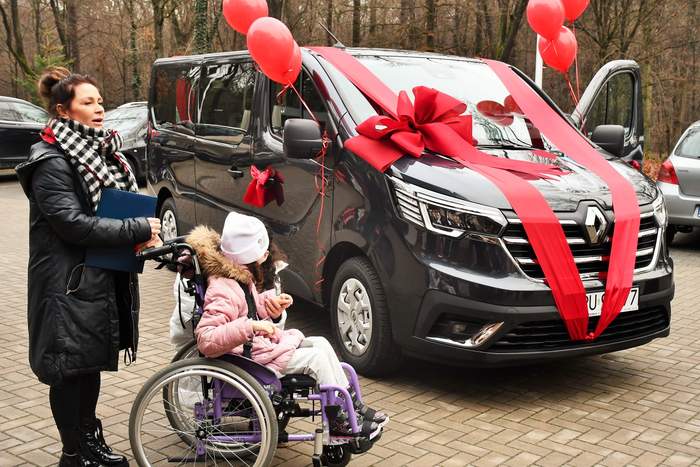 Pokaż zdjęcie: Na zdjęciu samochód bus marki Renault z przypiętymi szarfami i balonami w kolorze czerwonym. Przy busie stoi matka z dzieckiem niepełnosprawnym siedzącym na wózku