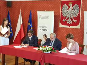 Pokaż zdjęcie: Trzy osoby siedzą przy stole konferencyjnym. Dwie z nich podpisuja dokumenty. Z tyłu kamerzysta filmuje podpisywanie umów