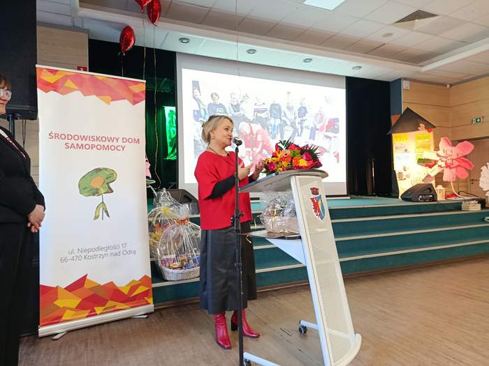 Pokaż zdjęcie: Dyrektor Elżbieta Miernik-Krukurka stoi z bukietem kwiatów przy pulpicie na tle banerów z logo ŚDS i Miasta Kostrzyn nad Odrą