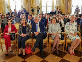 Pokaż zdjęcie: Na pierwszym planie pięć osób siedzi na fotelach w eleganckiej sali. Za nimi znajduje sie kilkadziesiąt siedzących, elegancko ubranych osób. Wystrój sali jest w kolorze złotym.