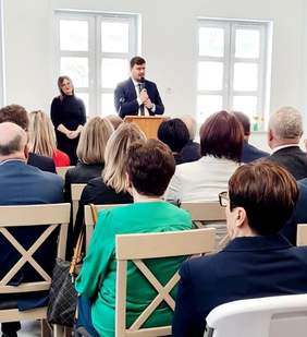 Pokaż zdjęcie: Mężczyzna w garniturze z mikrofonem przemawia. Przed nim kilkanaście osób siedzących na jasnych krzesłach
