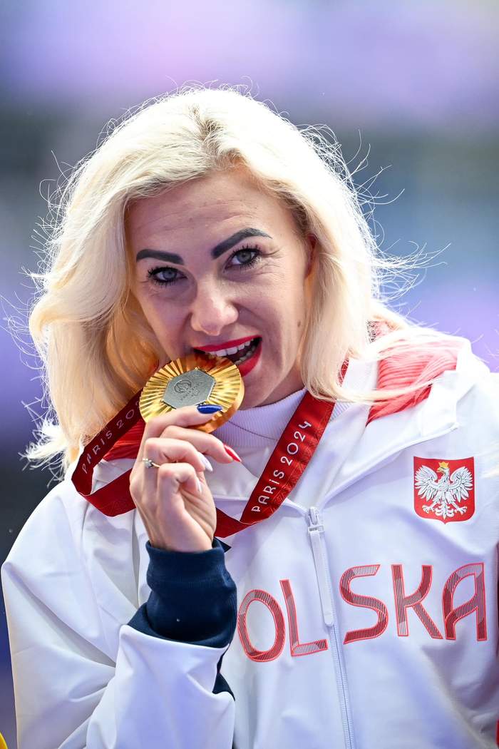 Pokaż zdjęcie: Kobieta gryzie złoty medal, jest ubrana w biały dres