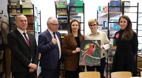 Pokaż zdjęcie: Grupa osób stojących w szkolnej bibliotece, jedna z nich trzyma pracę plastyczną przedstawiającą kolorowego ptaka.
