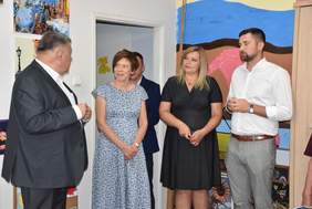 Pokaż zdjęcie: Dwie kobiety oraz trzech mężczyzn stoją w sali, w której znajduje się dużo kolorowych prac plastycznych. Jeden z mężczyzn przemawia w danym momencie, gdyż możemy dostrzec, że oczy pozostałych zgromadzonych osób są na niego skierowane.