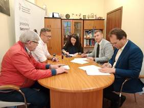 Pokaż zdjęcie: Przy owalnym stole siedzi kobieta i czterech mężczyzn. Podpisują umowę. Na stole leżą różne dokumenty, telefony, pieczątki i długopisy. 
