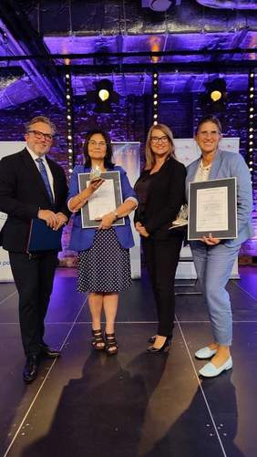 Pokaż zdjęcie: Stoją cztery osoby: trzy kobiety i jeden mężczyzna. Dwie panie prezentują otrzymane dyplomy i statuetki. Ściany wnętrza są fioletowe. 