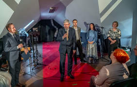 Pokaż zdjęcie: Na scenie stoi dyrektor Łódzkiego Oddziału PFRON, który przemawia do mikrofonu. Wokół niego znajdują sie inni ludzi. Widać także osoby siedzące na widowni.  