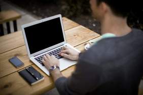 Pokaż zdjęcie: Mężczyzna pracuje przy komputerze, który leży na biurku. Obok laptopa znajduje się notes, długopis, telefon i filiżanka z kawą. 