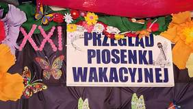 Pokaż zdjęcie: Na ścianie znajduje się plakat z napisem: Przegląd Piosenki Wakacyjnej. Wokół niego są różne dekoracje z kolorowego papieru i bibuły: motylki, kwiatuszki. 