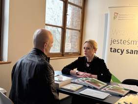 Pokaż zdjęcie: Mężczyzna i kobieta siedzą przy stole, rozmawiają. Na stole leżą broszury i dokumenty, a w tle znajduje się baner Państwowego Funduszu Rehabilitacji Osób Niepełnosprawnych z hasłem: „Wszyscy jesteśmy tacy sami.”