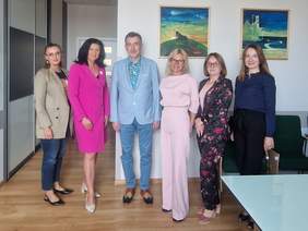 Pokaż zdjęcie: Sześć osób stoi w sali konferencyjnej na tle kolorowych obrazów; w środku mężczyzna w błękitnej marynarce, obok niego kobiety w eleganckich strojach.
