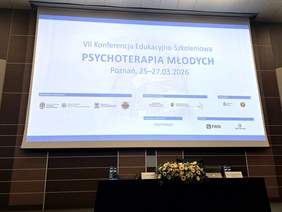 Pokaż zdjęcie: Ekran projekcyjny w sali konferencyjnej z wyświetloną planszą wydarzenia. Na środku slajdu jest duży tytuł, a pod nim data i miejsce. Na dole znajdują się logotypy partnerów. Pod ekranem stoi długi stół, a na nim w centrum kompozycja kwiatowa. Po bokach widać drobny sprzęt nagłośnieniowy. Tekst widoczny na slajdzie: VII Konferencja Edukacyjno-Szkoleniowa, a pod nim PSYCHOTERAPIA MŁODYCH oraz data i miejsce wydarzenia: Poznań, od 25 do 27 marca 2026 roku