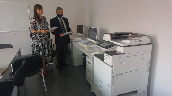 Pokaż zdjęcie: W pomieszczeniu pomalowanym na biało stoi kobieta i mężczyzna, oboje trzymają w rękach kartki papieru. Stoją pomiędzy stołem, krzesłami i dwoma białymi biurkami na których znajdują się monitory i dokumenty. przy pierwszym biurku stoi urządze