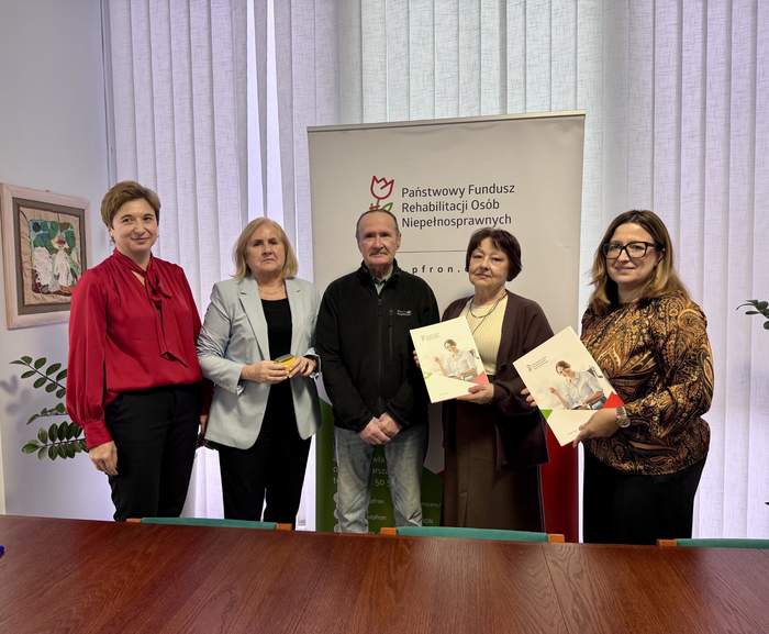 Pokaż zdjęcie: Na tle rollupu z logo PFRON pozują cztery kobiety i jeden mężczyzna, dwie kobiety trzymają w rękach umowę.