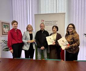 Pokaż zdjęcie: Na tle rollupu z logo PFRON pozują cztery kobiety i jeden mężczyzna, dwie kobiety trzymają w rękach umowę.