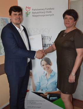 Pokaż zdjęcie: Kobieta i mężczyzna podają sobie dłoń. Stoją na tle grafiki PFRON