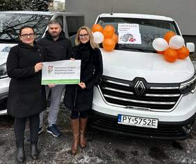 Pokaż zdjęcie: Dwie kobiety trzymają w dłoniach naklejkę z logo PFRON informującym o dofinansowaniu ze środków PFRON. Z tyłu stoi mężczyzna. W tle dwa busy. Jeden z nich udekorowany balonami. Na szybie pojazdu naklejka z logo Przedszkola Specjalnego „Orzeszek”.