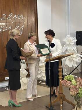 Pokaż zdjęcie: Trzy kobiety stojące obok siebie podczas uroczystości. Dyrektor Oddziału Podlaskiego PFRON wręcza dyrektor DPS w Brańsku grawerton i kwiaty. Obok stoi przedstawicielka PFRON trzymająca bukiet.