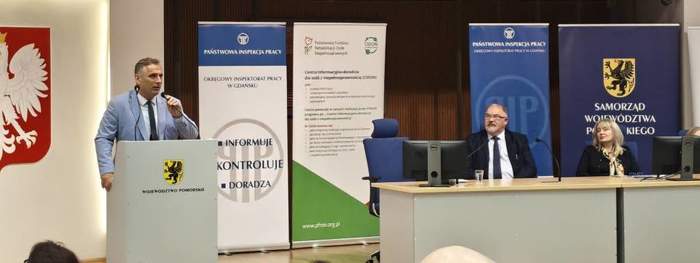 Pokaż zdjęcie: Mężczyzna w jasnym garniturze przemawia z mównicy podczas konferencji w sali urzędu. Na scenie siedzą dwie osoby przy stole z mikrofonem. W tle widoczne są banery Państwowej Inspekcji Pracy i Samorządu Województwa Pomorskiego.