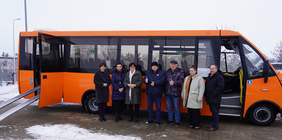 Pokaż zdjęcie: Pomarańczowy autobus stojący na zewnątrz, na tle zimowego krajobrazu.   Przed boczną ścianą autobusu stoi w szeregu grupa siedmiu osób w zimowych ubraniach – płaszczach i kurtkach, w ciemnych i jasnych kolorach.   Autobus jest średniej wielkości, ma prostokątne okna wzdłuż boku i otwarte przednie drzwi z prawej i lewej strony. Przy drzwiach z lewej strony ustawione są najazdy aby można było wjechać wózkiem. 