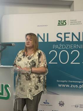 Pokaż zdjęcie: Osoba stojąca przy mównicy z napisem ZUS. W tle baner informujący o dniach seniora.
