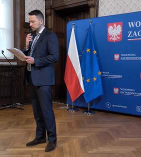 Pokaż zdjęcie: Dyrektor Oddziału w granatowym garniturze na tle napisu Województwo Zachodniopomorskie mówi do mikrofony trzymając kartkę w ręku.