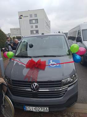Pokaż zdjęcie: Przód samochodu udekorowany jest czerwoną kokardką i kolorowymi balonami. Po lewej stronie widać fragment wózka inwalidzkiego. W tle znajduje się operator z kamerą. Na aucie naklejona jest naklejka symbolizująca przewóz osób z niepełnosprawnością. 