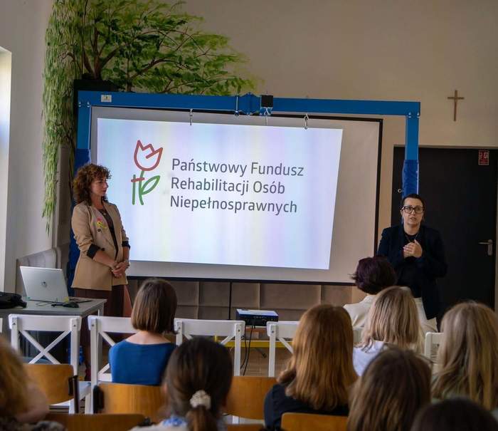 Pokaż zdjęcie: Prezentacja PFRON podczas spotkania „Spektrum Możliwości” w szkole w Lublinie; dwie osoby prowadzą wystąpienie przed uczestnikami, na ekranie widoczne logo PFRON.