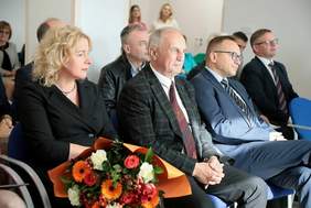 Pokaż zdjęcie: Zdjęcie osob siedzących w sali na otwarciu zakładu opiekuńczo mieszkalnego, zbliżenie na kobietę blondynkę w czarnej sukience oraz dwóch mężczyzn w garniturze