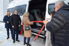 Pokaż zdjęcie: Przy białym autobusie, stoja cztery osoby, trzech mężczyzn i jedna kobieta. Kobieta blondynka w beżowym płaszczu przecina nożyczkami czerwoną wstęgę
