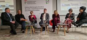Pokaż zdjęcie: Paneliści kobiety i mężczyźni siedzą na środku sali na krzesłach. Dyskusję moderuje niewidomy mężczyzna - Marcin Ryszka.
