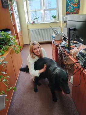 Pokaż zdjęcie: Na korytarzu stoi duży, czarny pies. Obok niego kuca kobieta, która go głaszcze. Pani się uśmiecha. 