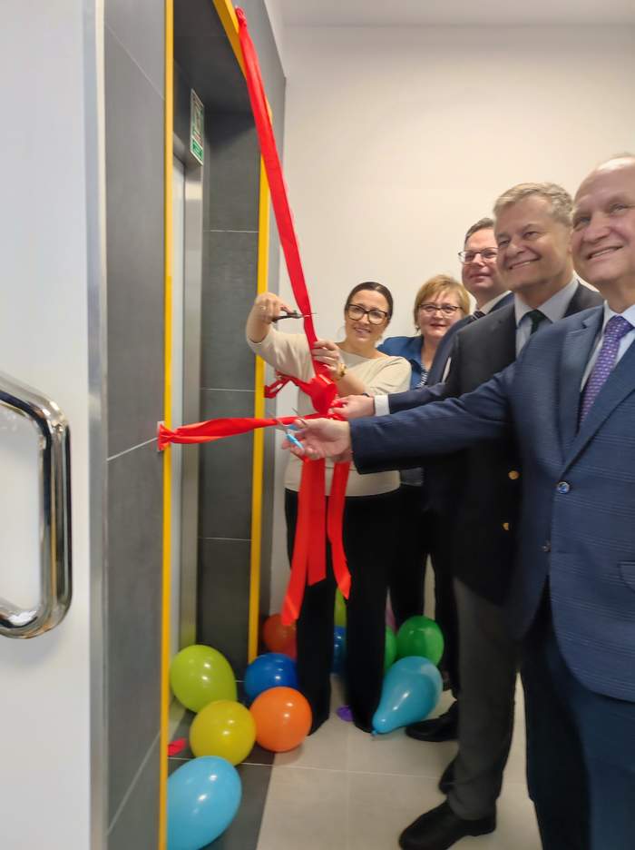 Pokaż zdjęcie: Grupa osób uroczyście przecina czerwoną wstęgę przed wejściem do nowej windy, otoczoną kolorowymi balonami, symbolicznie inaugurując nową, dostępną przestrzeń w budynku.