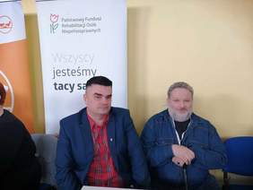 Pokaż zdjęcie: Dwóch mężczyzn na krzesłach, za nimi baner Państwowego Funduszu Rehabilitacji Osób Niepełnosprawnych z napisem ,,Wszyscy jesteśmy tacy sami’'