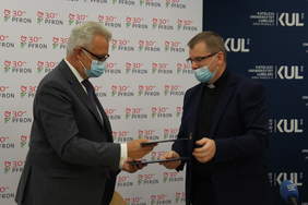 Pokaż zdjęcie: Mężczyzna w garniturze uroczyście wręczający księdzu, dwie granatowe teczki. Obaj w maseczkach higienicznych. Na pierwszym planie mikrofon Polskiego Radia Lublin. W tle bannery Państwowego Funduszu Rehabilitacji Osób Niepełnosprawnych i Katolicki