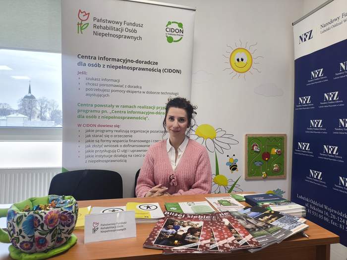 Pokaż zdjęcie: Stoisko informacyjne CIDON z materiałami promocyjnymi na biurku, przedstawicielka organizacji siedzi przy stole. W tle banery PFRON i NFZ oraz kolorowe ilustracje.